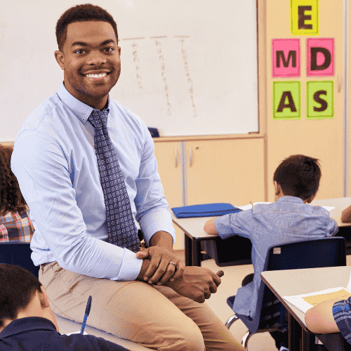 Photo of a male teacher sitting on a desk in a classroom.